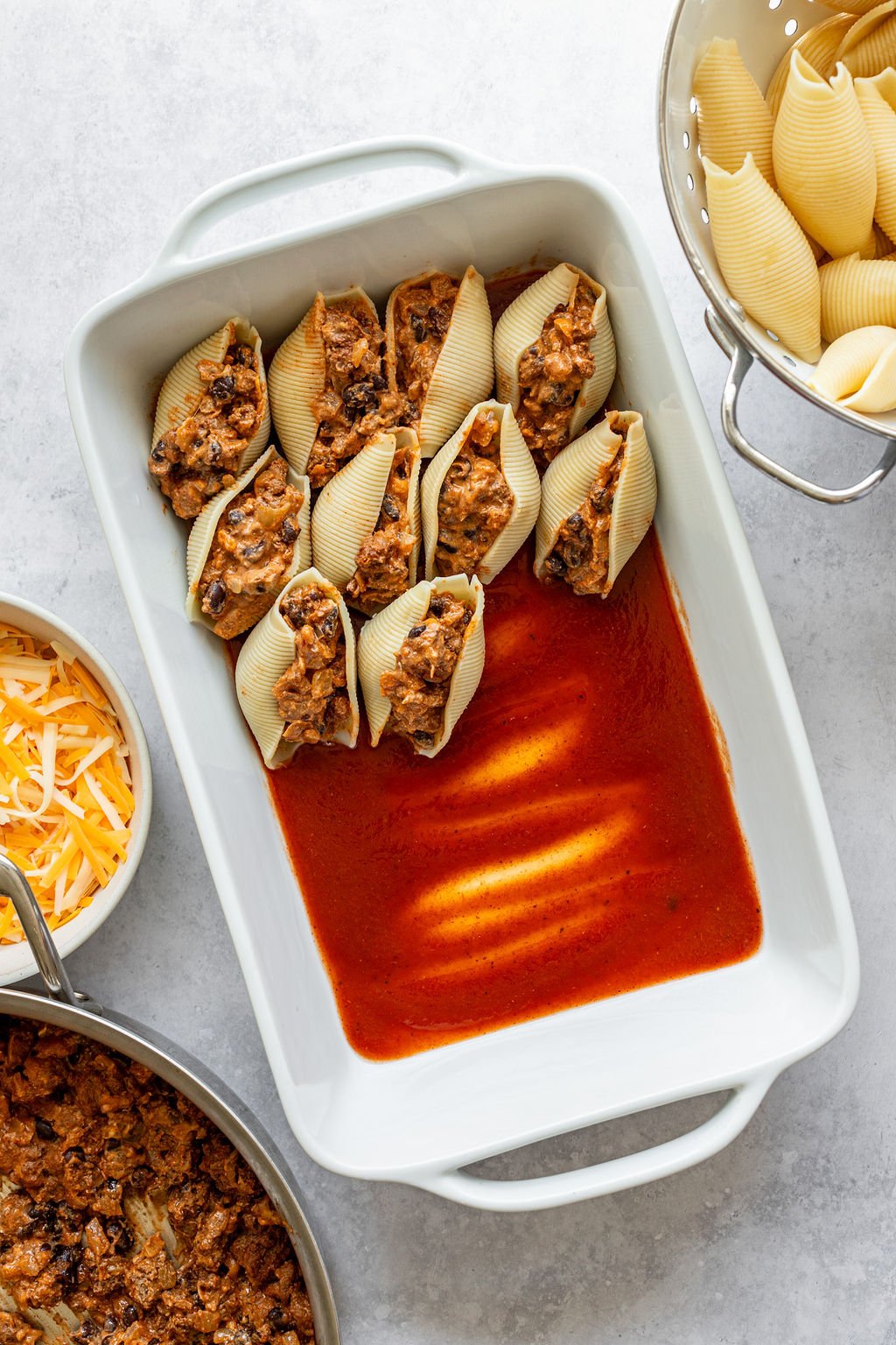 Baking dish with red sauce and pasta shells filled with ground beef and cheese. Baking dish is surrounded by a bowl of shredded cheese, colander of cooked pasta shells, and saucepan of cooked ground beef.