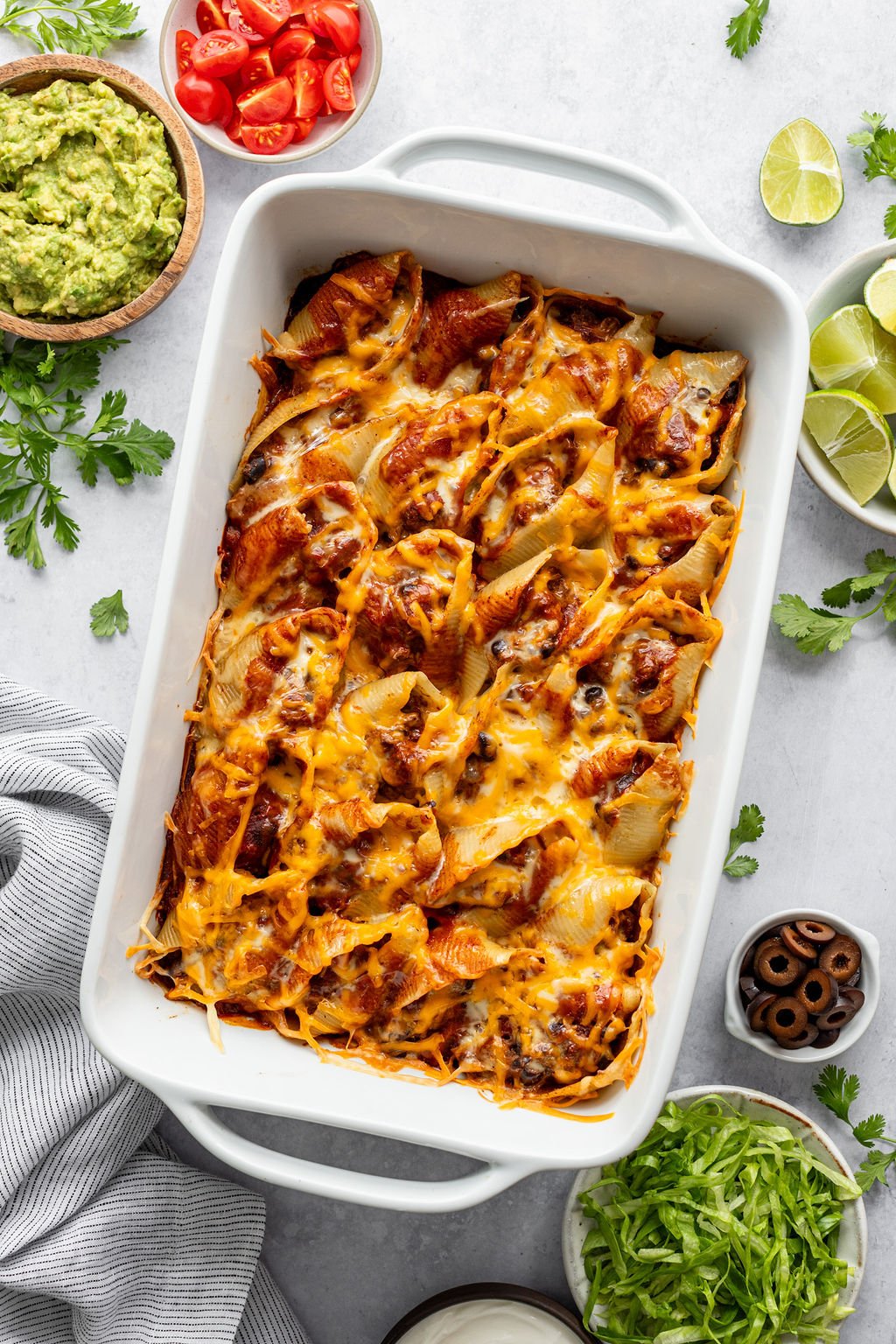 Baking dish with pasta shells filled with ground beef and topped with red sauce and melted cheese. Baking dish is surrounded by a dish towel and small bowls of guacamole, diced tomatoes, lime wedges, shredded lettuce, and sliced olives.