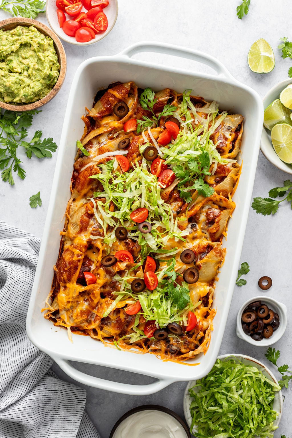 Baking dish with pasta shells filled with ground beef and topped with red sauce, melted cheese, shredded lettuce, diced tomatoes, cilantro, and olives. Baking dish is surrounded by a dish towel and small bowls of guacamole, diced tomatoes, lime wedges, sour cream, shredded lettuce, and sliced olives.