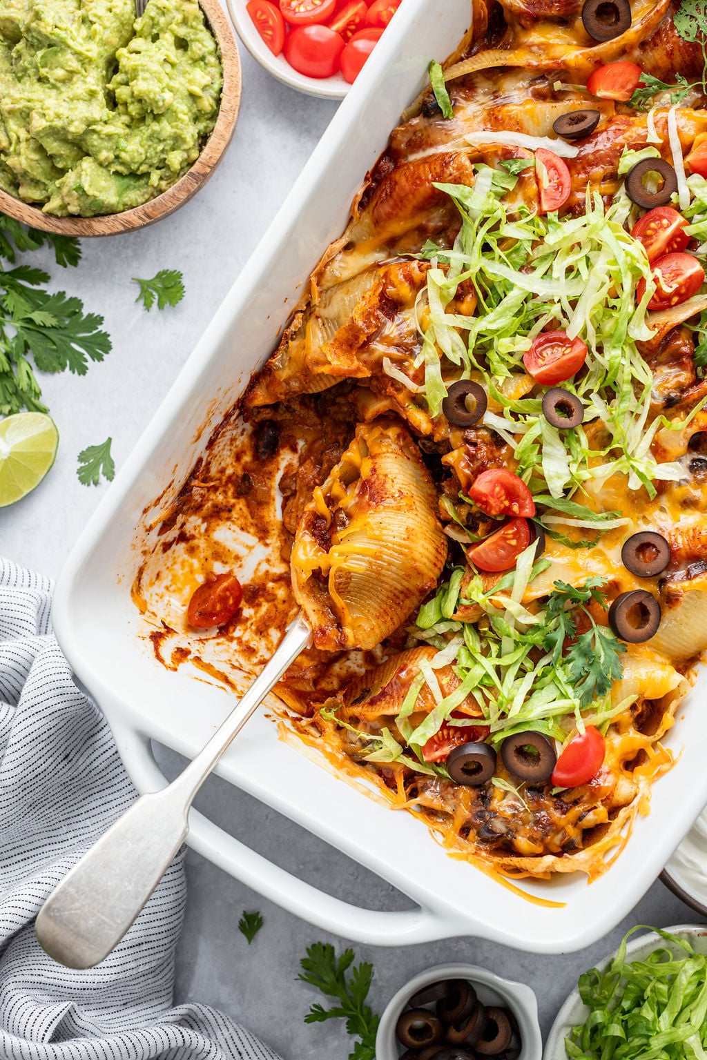 A baking dish of stuffed pasta shells topped with melted cheese, lettuce, tomatoes, and olives, with a serving spoon and sides of guacamole and chopped tomatoes nearby.