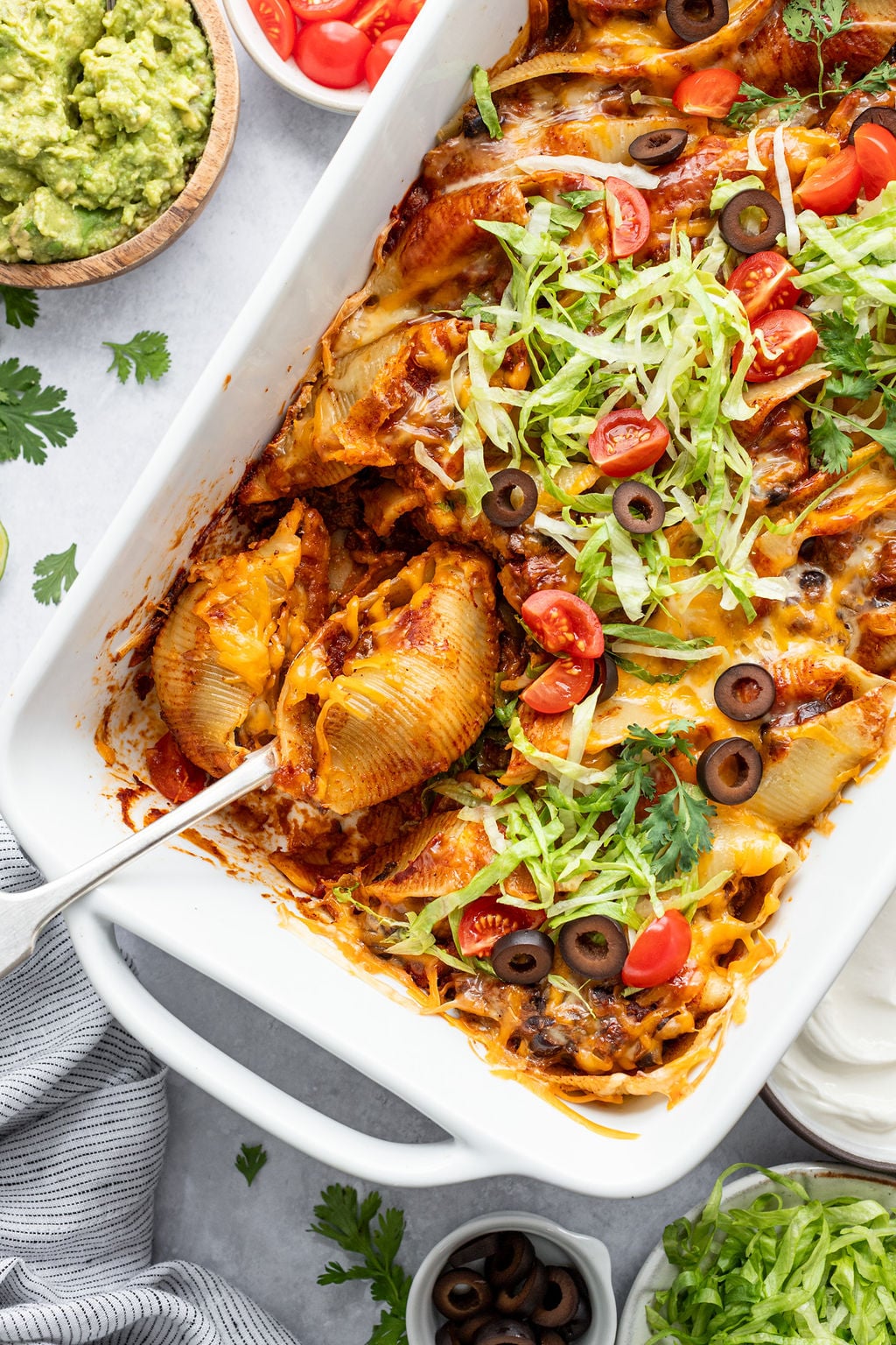 Close-up of a baking dish with a spoon and pasta shells filled with ground beef and topped with red sauce, melted cheese, shredded lettuce, diced tomatoes, cilantro, and olives. Baking dish is surrounded by a dish towel and small bowls of guacamole, diced tomatoes, shredded lettuce, sour cream, and sliced olives.