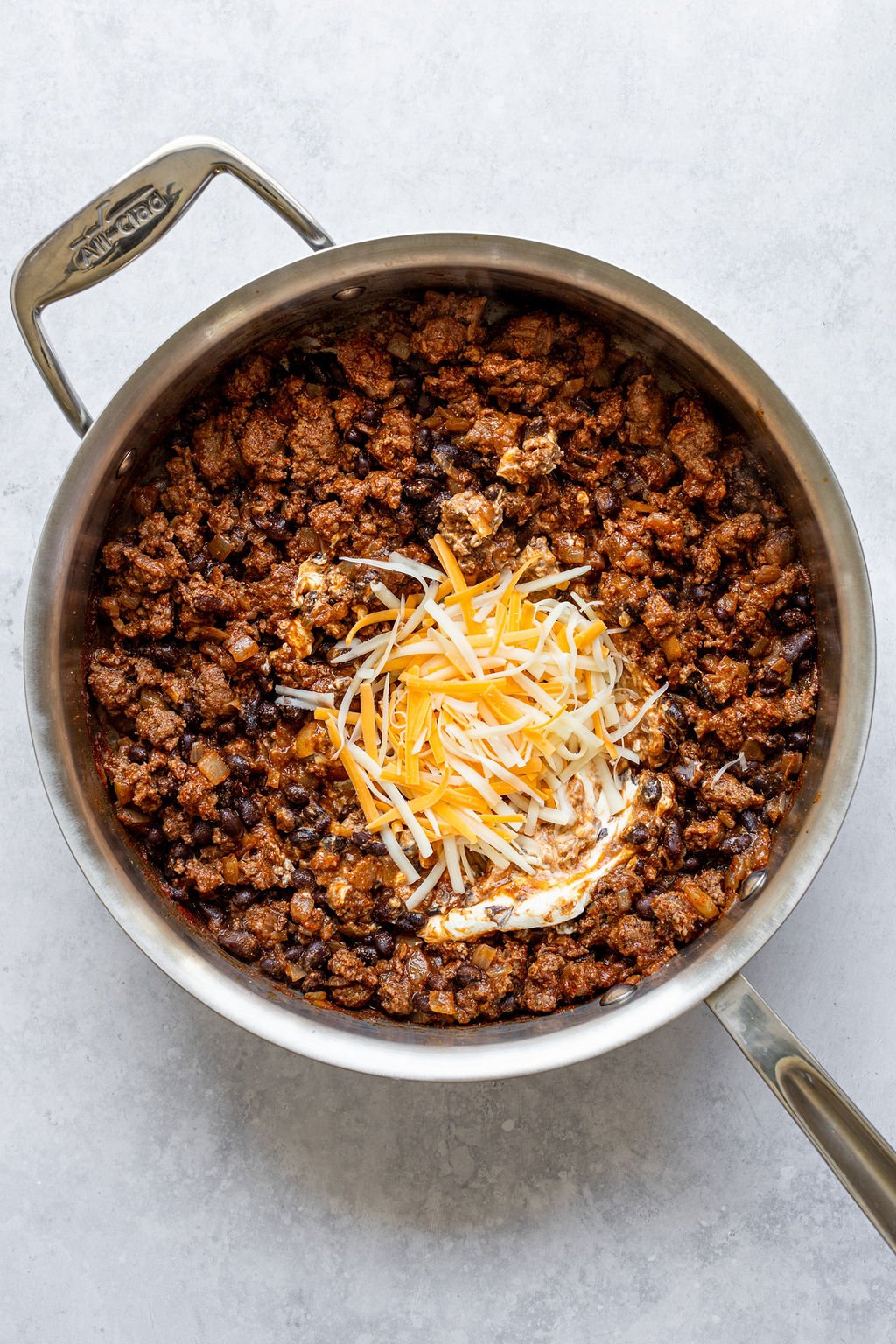 Saucepan with cooked ground beef and black beans, topped with shredded cheese.