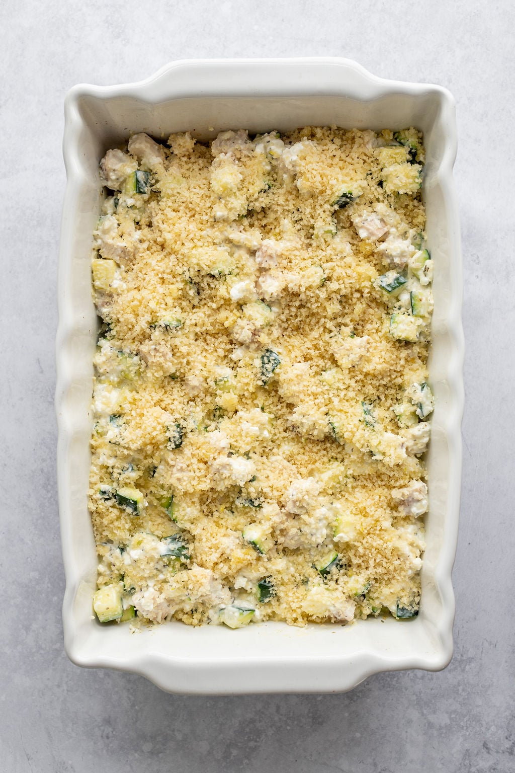 White baking dish with diced zucchini and diced chicken in a creamy sauce and topped with breadcrumbs.