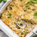 White baking dish on top of a towel with a spoon and a baked zucchini casserole covered in breadcrumbs and topped with basil.