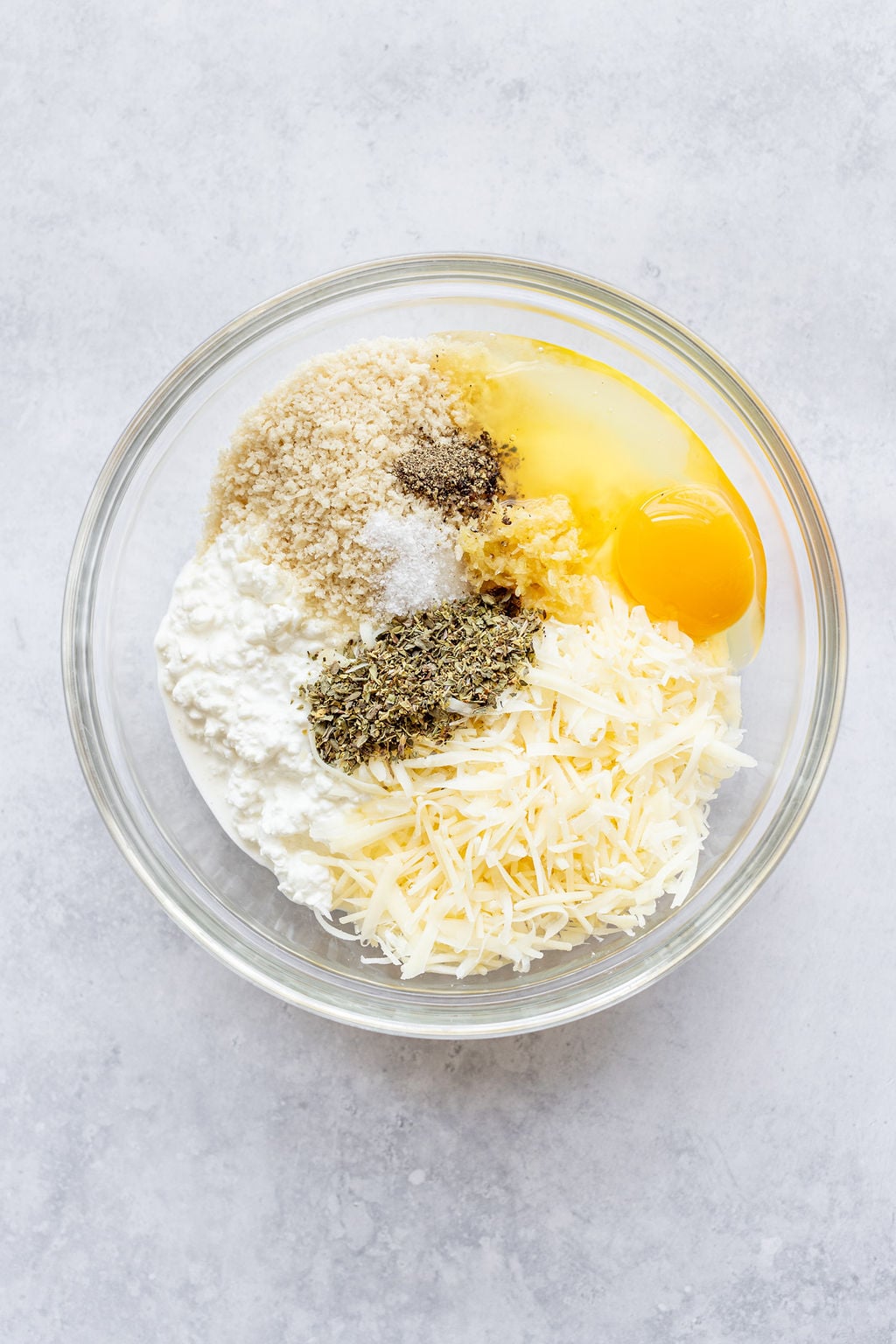Glass bowl with breadcrumbs, cottage cheese, shredded cheese, herbs, pepper, salt, and an egg.