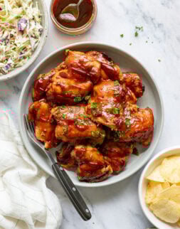 Overhead shot of a serving bowl with a serving fork and cooked chicken thighs covered in red sauce and chopped fresh herbs. Bowl is surrounded by a towel; a jar of red sauce; and small bowls of coleslaw, potato chips, and chopped herbs.