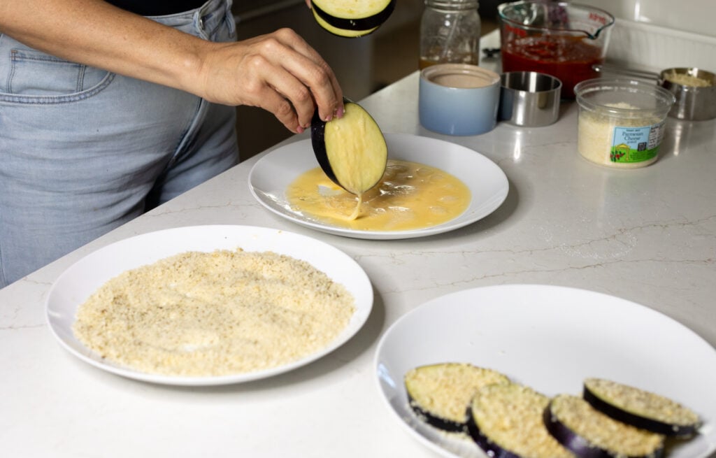 White plates with a yellow liquid, breadcrumbs, and eggplant slices covered in breadcrumbs. Someone is dipping eggplant slices in the yellow liquid.
