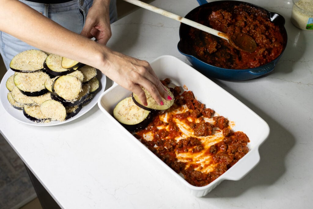 A skillet with a red meat sauce, a plate with breaded eggplant slices, and a white baking dish with red meat sauce and eggplant slices. Someone is placing eggplant slices into the baking dish.