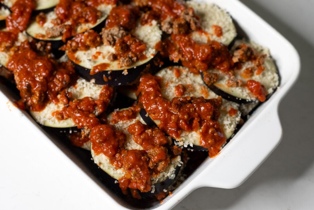 Close-up of a white baking dish with eggplant slices covered in a red meat sauce.