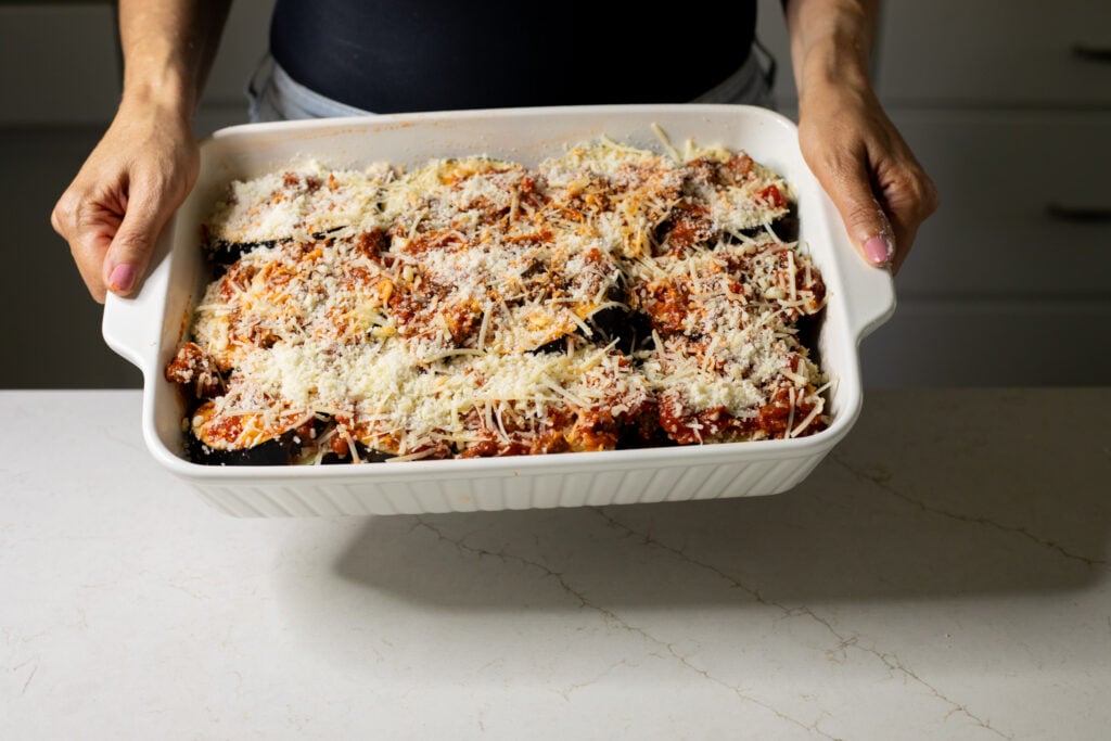 Someone holding a white baking dish with eggplant slices covered in a red sauce and topped with shredded cheese.
