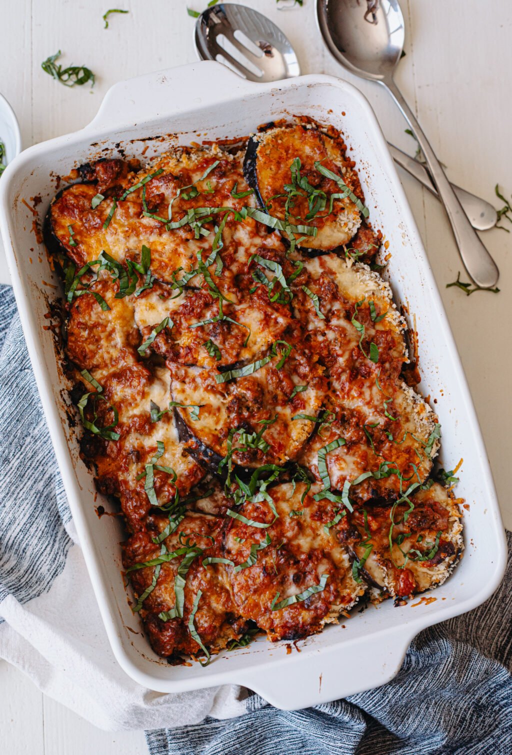 Close-up of a white baking dish with eggplant slices topped with cheese, tomato sauce, and fresh basil. Baking dish is surrounded by a dish towel, serving spoons, and a small bowl of fresh basil.