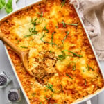 A baked beef and zucchini casserole in a white dish, topped with melted cheese and fresh basil, with a wooden spoon serving a portion.