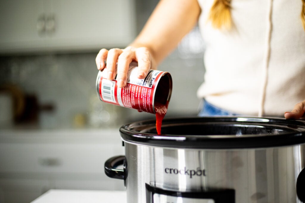 Person adding tomato sauce to a slow cooker.