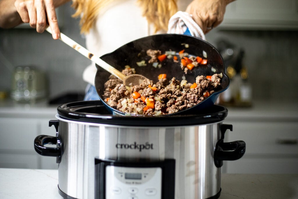 Person adding cooked ground beef and vegetables to a slow cooker.