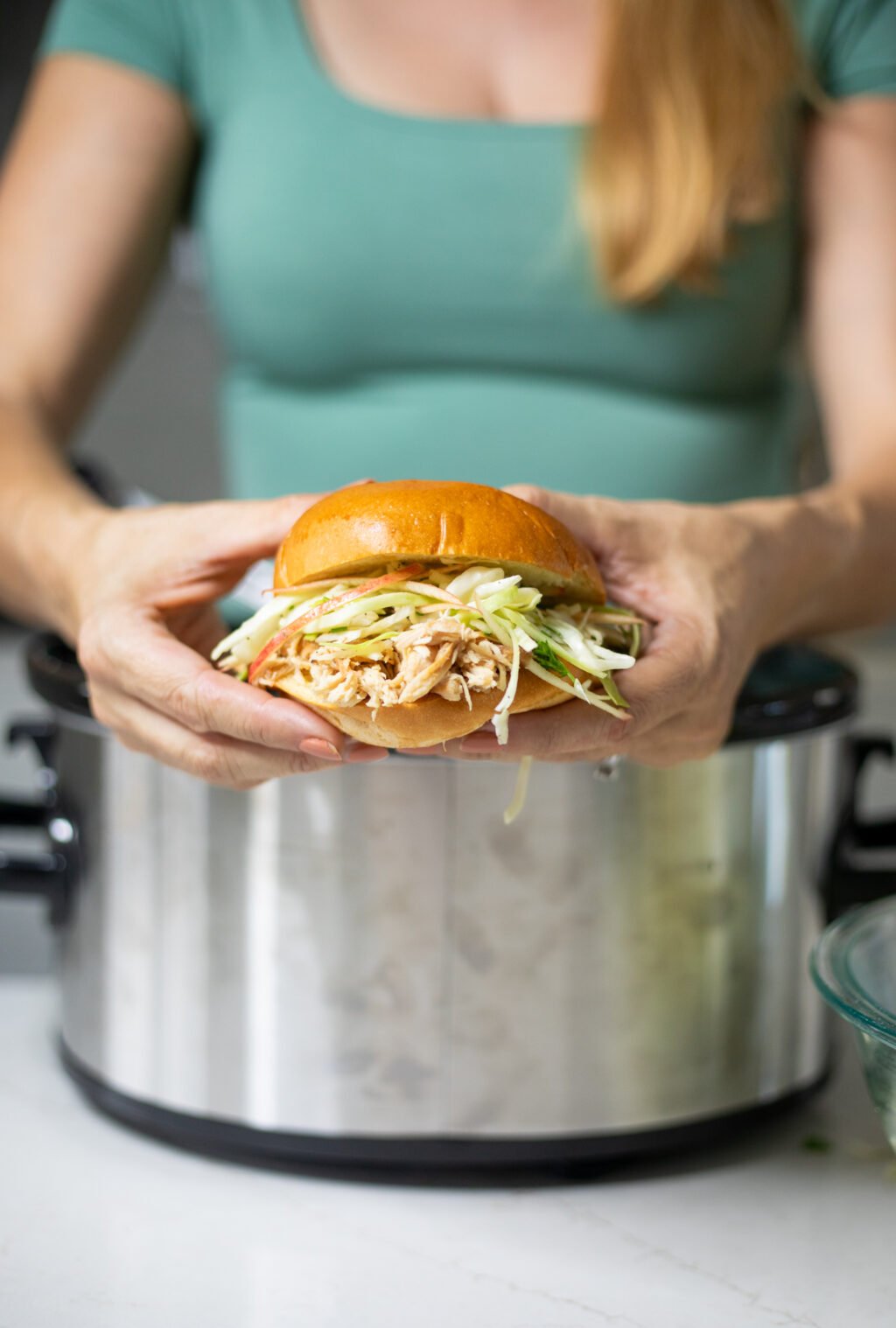 Person holding a bun filled with shredded chicken and shredded cabbage and apple slaw.