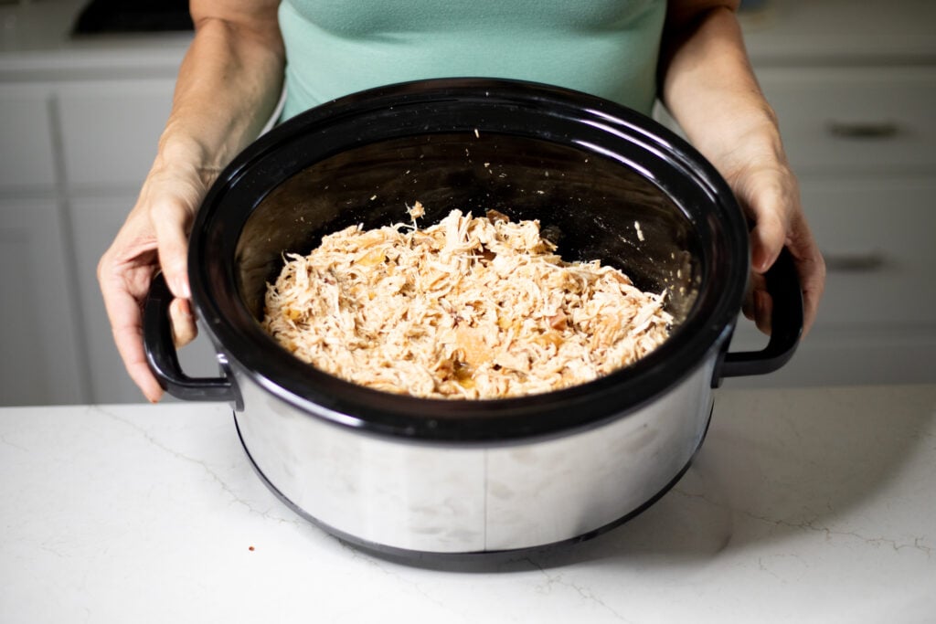 Person holding a slow cooker filled with shredded chicken.