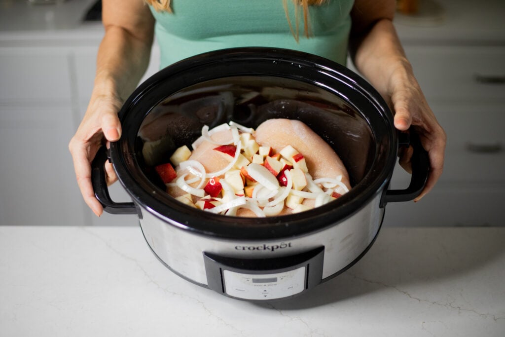 Person holding a slow cooker filled with raw chicken, sliced onion, and diced apple.