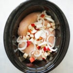 Overhead shot of a slow cooker filled with raw chicken, chopped apple, and sliced onion.