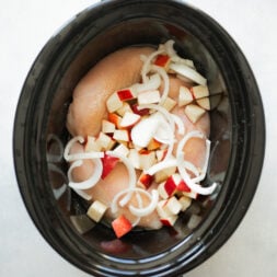 Overhead shot of a slow cooker filled with raw chicken, chopped apple, and sliced onion.