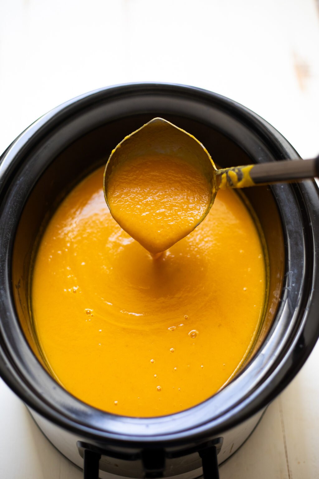 Ladle pouring yellow butternut squash soup into a black slow cooker.
