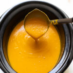 Ladle pouring yellow butternut squash soup into a black slow cooker.