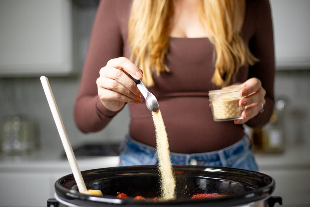Person pouring a spoonful of garlic powder into a slow cooker.