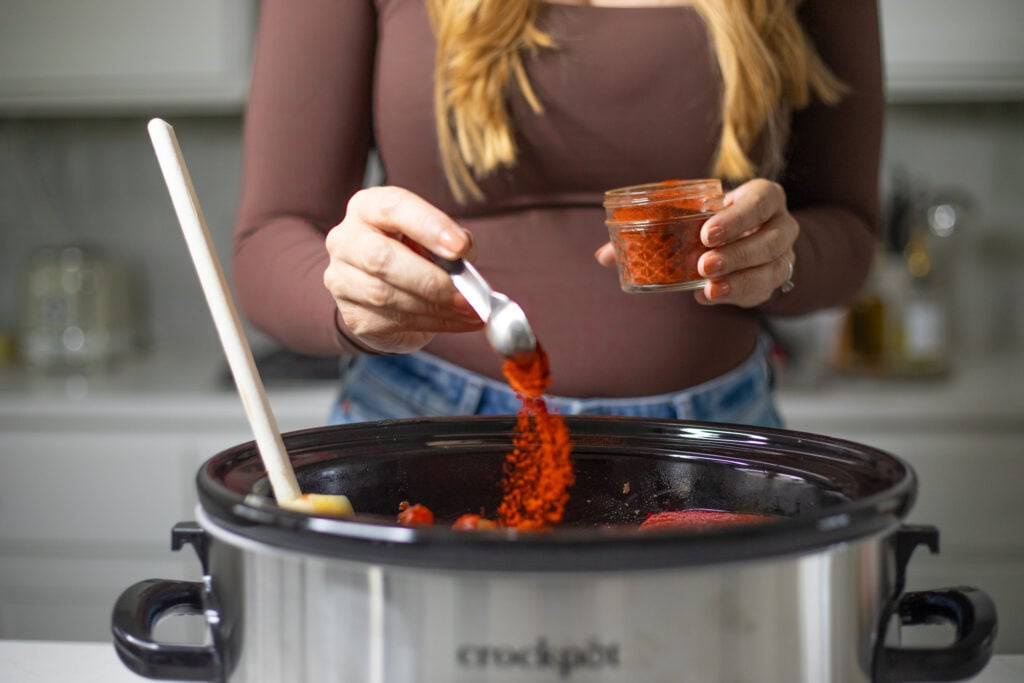 Person pouring red spice into slow cooker.