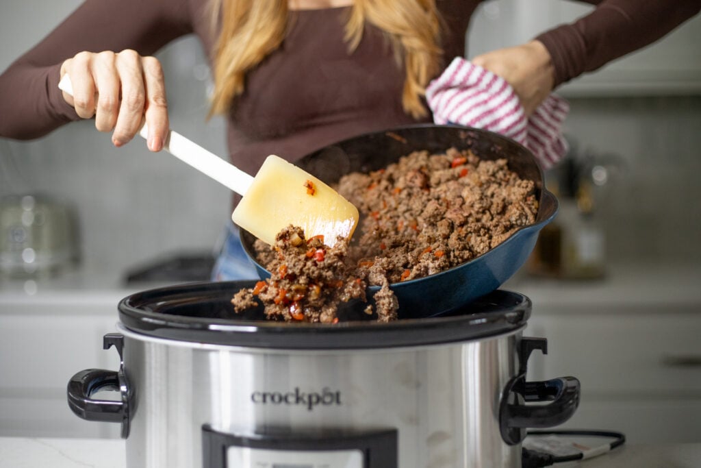 Person pouring cooked ground beef from a skillet into slow cooker.