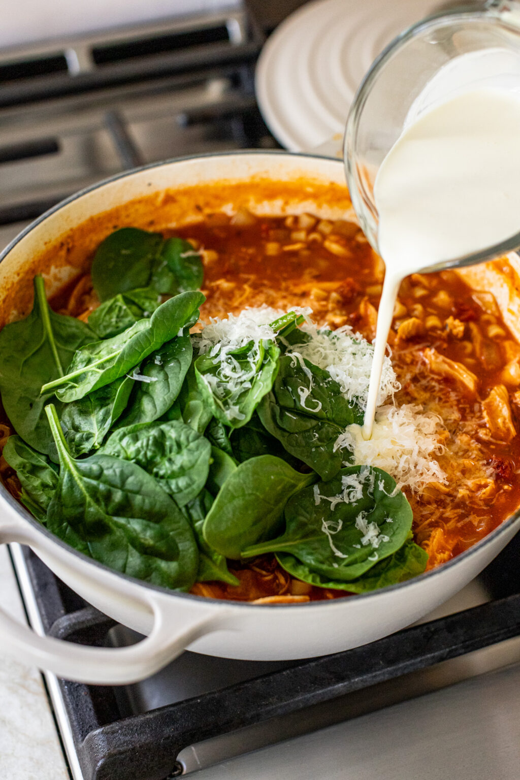 Cream being poured from a glass into a large pot of chicken tomato soup topped with spinach and shredded cheese. 