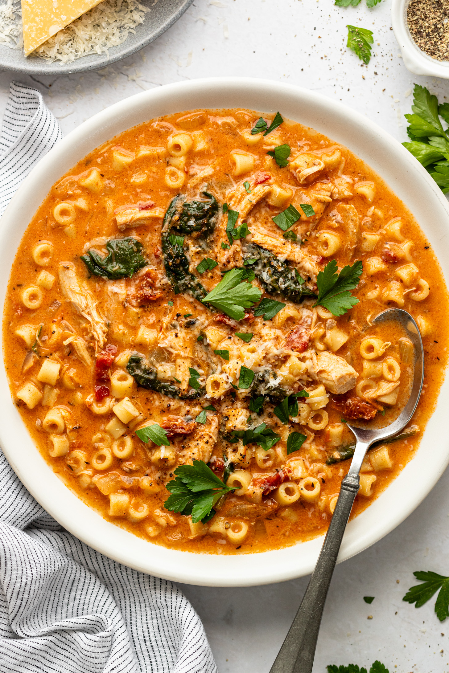 A metal spoon in a bowl of creamy tomato soup with small pasta, shredded chicken, spinach, sundried tomatoes, and fresh parsley. 