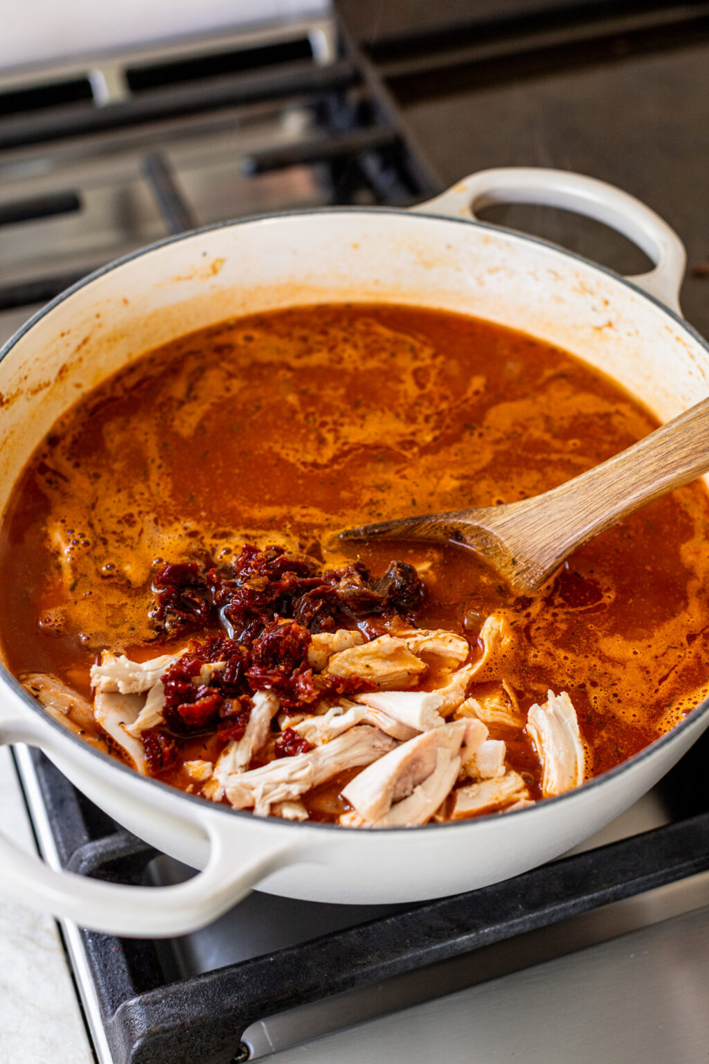 Shredded chicken and sun dried tomatoes sitting on top of tomato soup in a large pot.