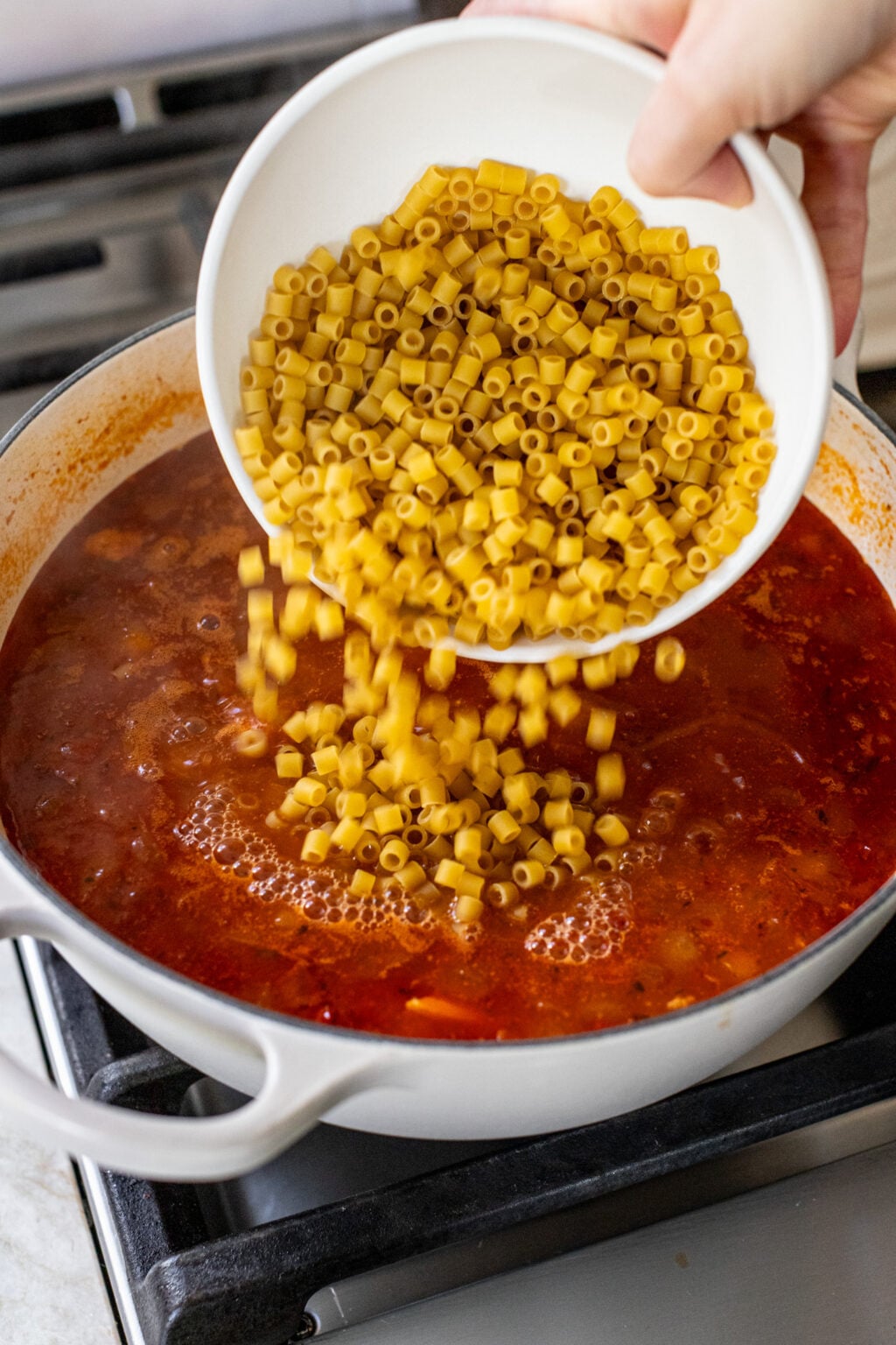Small pasta being poured from a bowl into a pot of creamy red tomato soup.