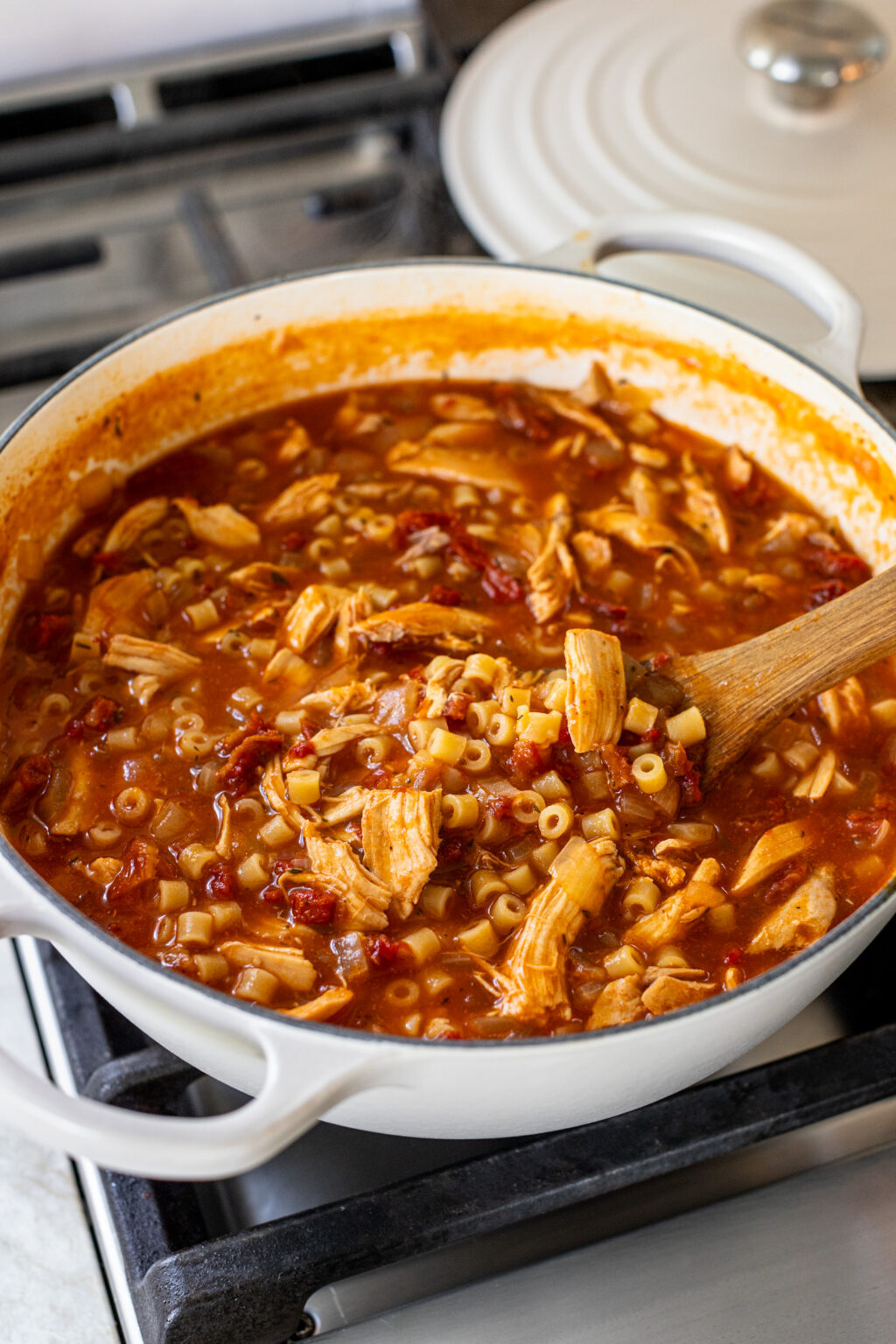 A spoon stirring a large pot of creamy red tomato soup with shredded chicken and small pasta. 