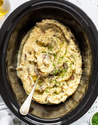 Creamy smashed potatoes in a large slow cooker with a large metal spoon.