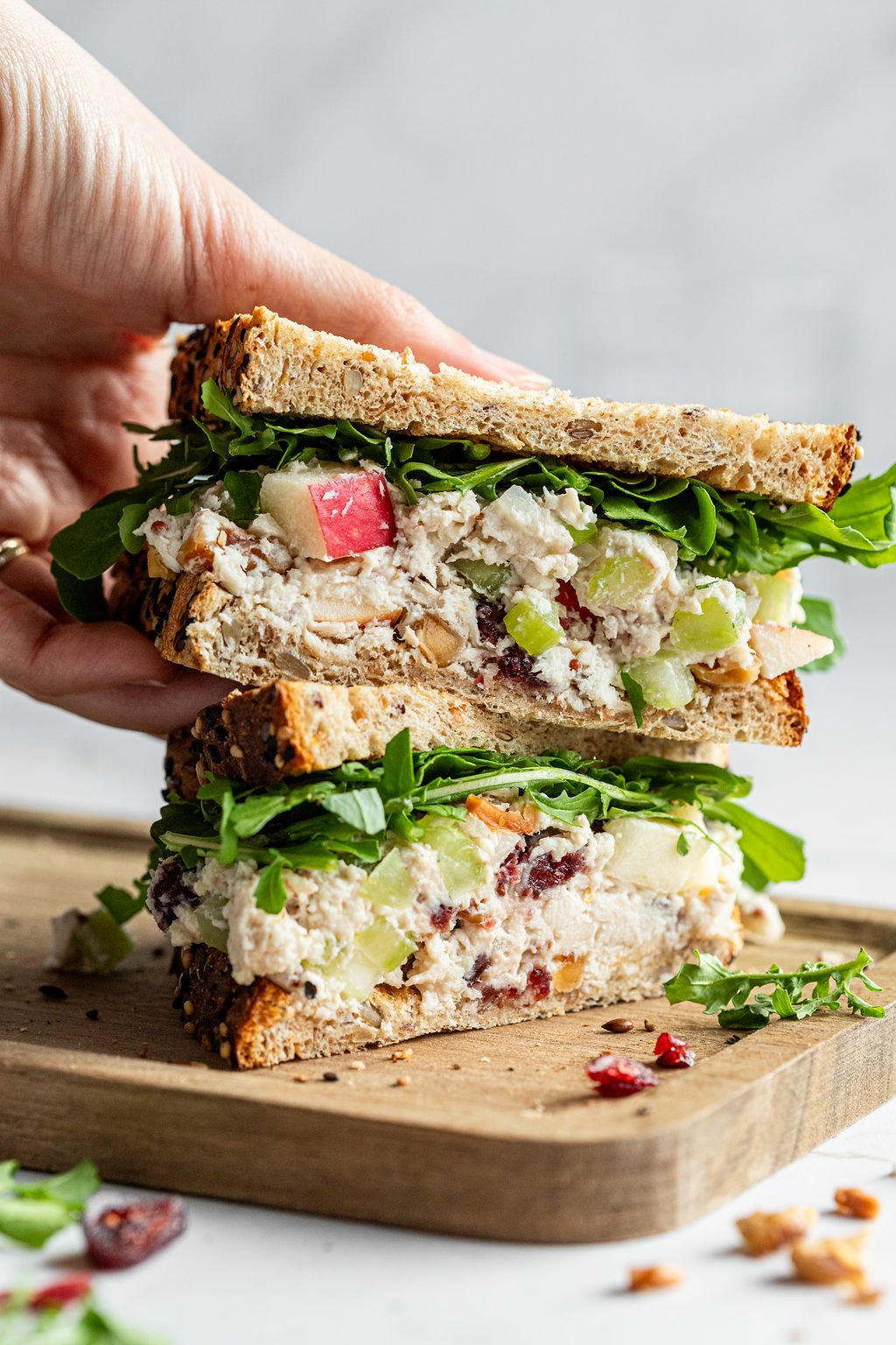 A hand lifting one half of a chicken salad sandwich off a wood cutting board.