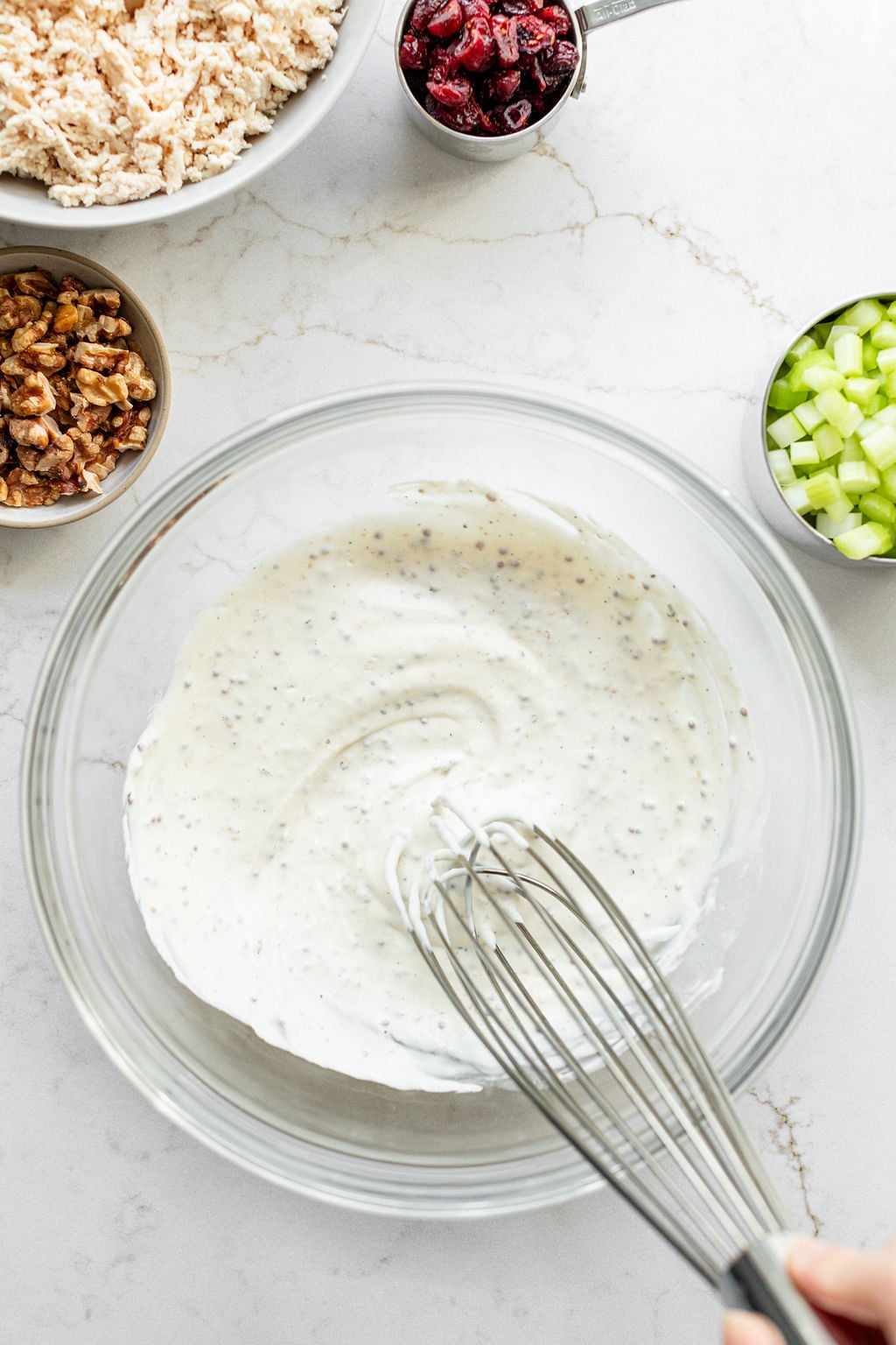 A hand whisking a light, creamy yogurt and mayonnaise mixture in a glass mixing bowl.