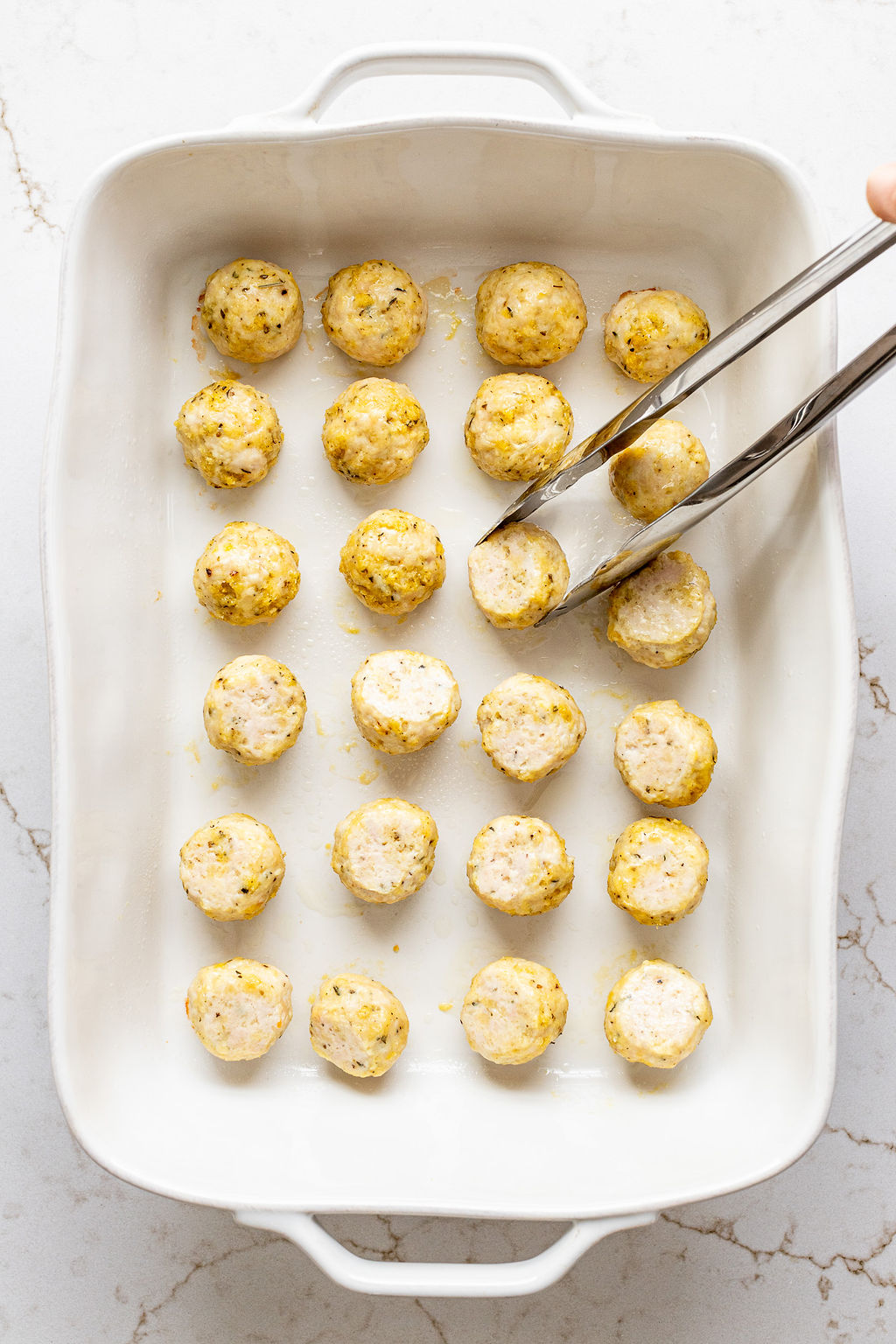 Metal tongs flipping chicken meatballs in a casserole dish.