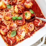 A metal spoon sitting in a large white baking dish filled with small chicken meatballs, creamy marinara sauce, and shredded Italian cheese.