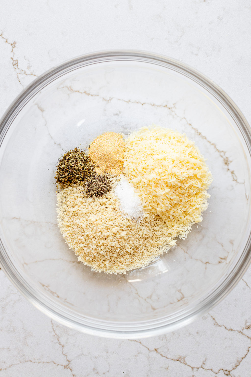 A glass mixing bowl filled with salt, pepper, garlic powder, bread crumbs, and parmesan cheese.