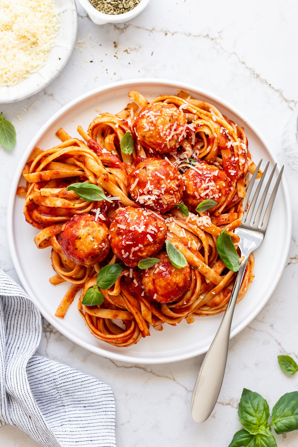 A plate of pasta covered in red sauce, grated cheese, and baked red meatballs with a fork.