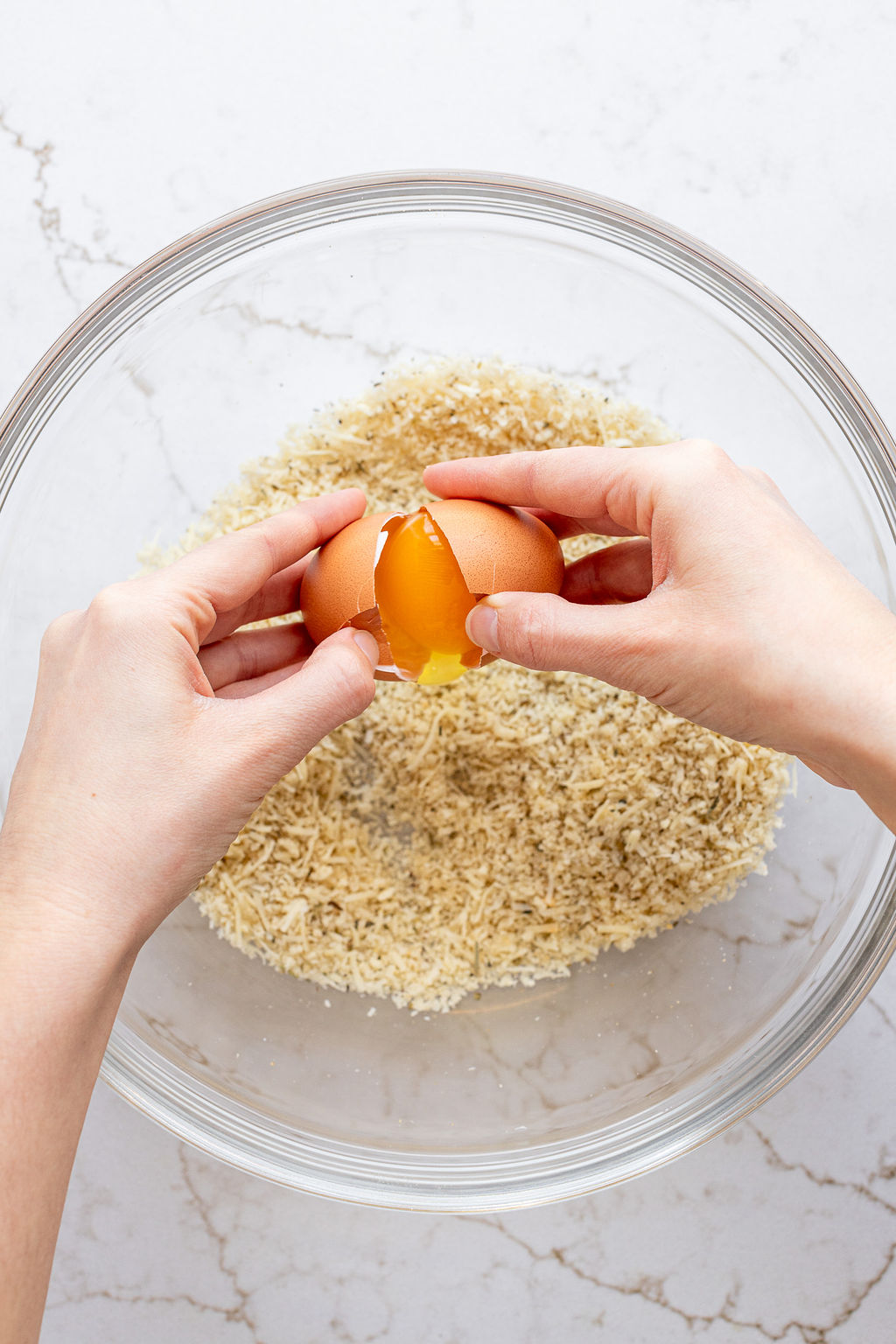 Two hands cracking an egg into a bowl with fine bread crumbs.