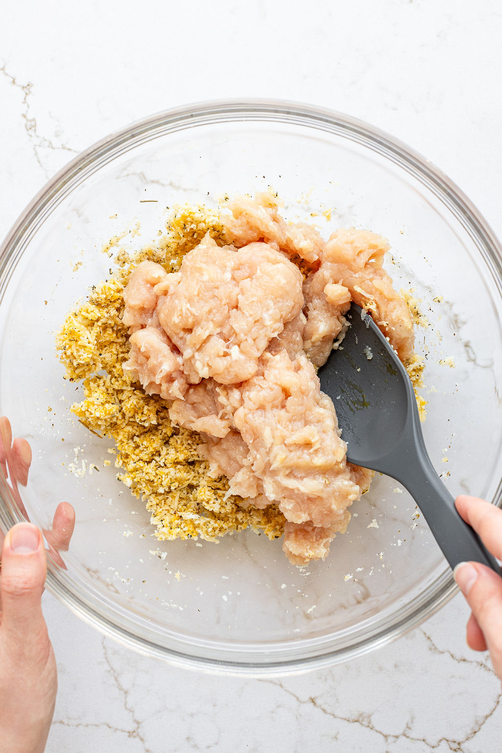 A hand mixing ground chicken with a spoon into a bowl of grated parmesan, bread crumbs, and seasonings.