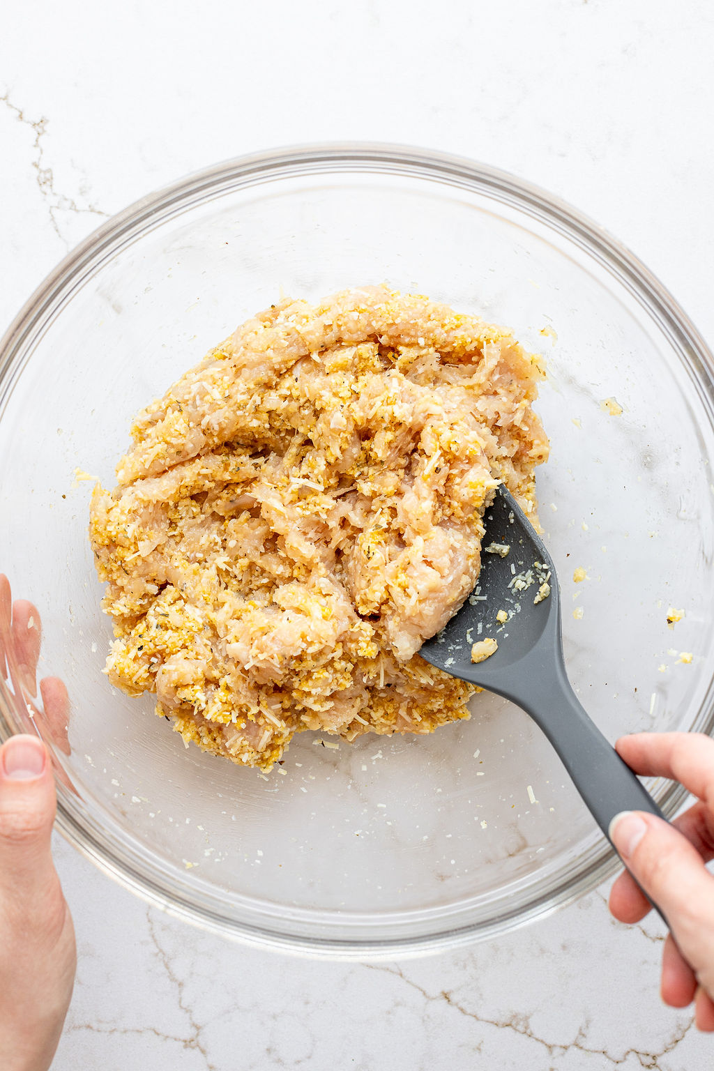 A hand mixing a sticky mixture of ground chicken, grated cheese, breadcrumbs, and seasonings with a large spoon.