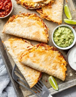 Five crispy, golden tacos on a baking sheet next to salsa and guacamole in bowls.