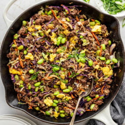 A large metal skillet filled with eggs, ground beef, coleslaw mix, and edamame on a marble surface.