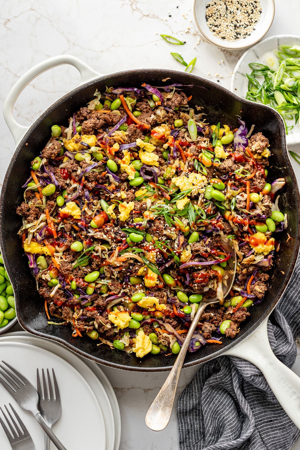 Cooked eggs, ground beef, coleslaw mix, and edamame in a large skillet with a spoon.