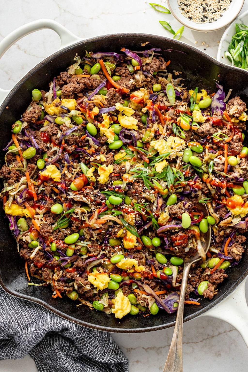 Sriracha and chopped green onions sprinkled over cooked beef, coleslaw, eggs, edamame, and brown sauce in a skillet.