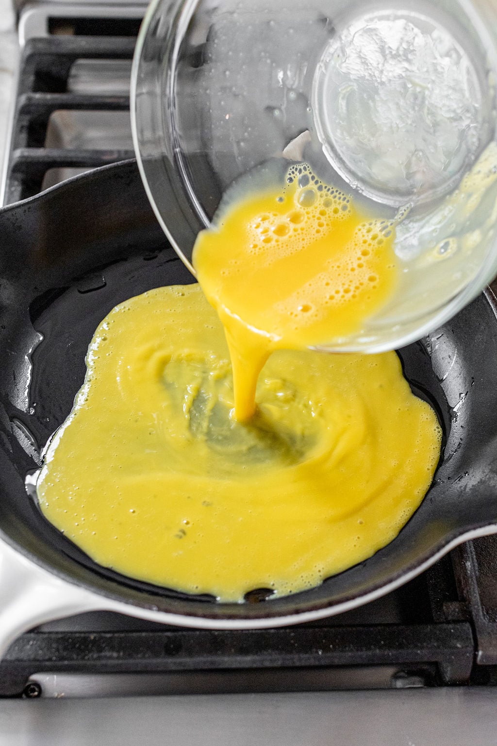 Whisked eggs being poured from a bowl into a large skillet on the stove.