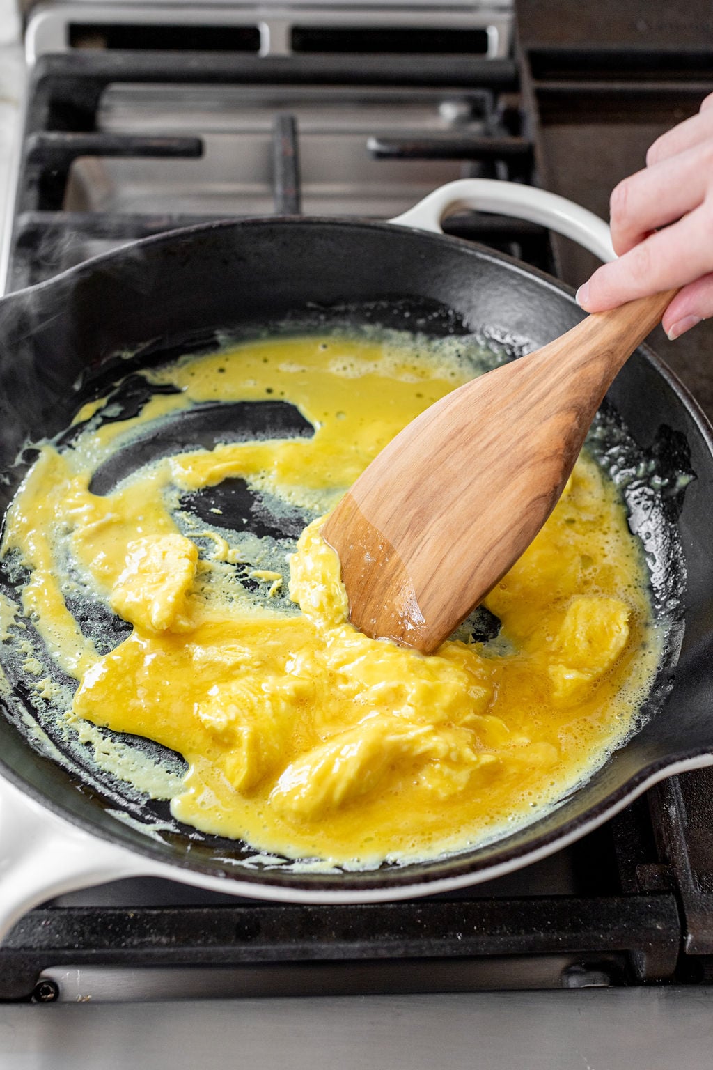 A hand stirring scrambled eggs in a large skillet on the stove.