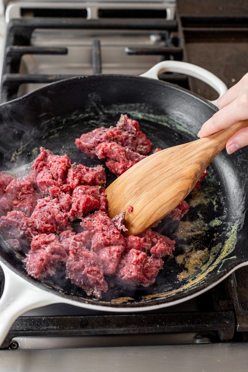 A hand using a large wooden spoon to break up ground beef in a skillet on the stovetop.